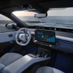 Wnętrze nowoczesnego samochodu BMW iX3 50 xDrive z wyświetlaczem multimedialnym i kierownicą o futurystycznym kształcie, z widokiem na morze w tle. Luksusowe detale i oświetlenie podkreślają premium charakter marki BMW