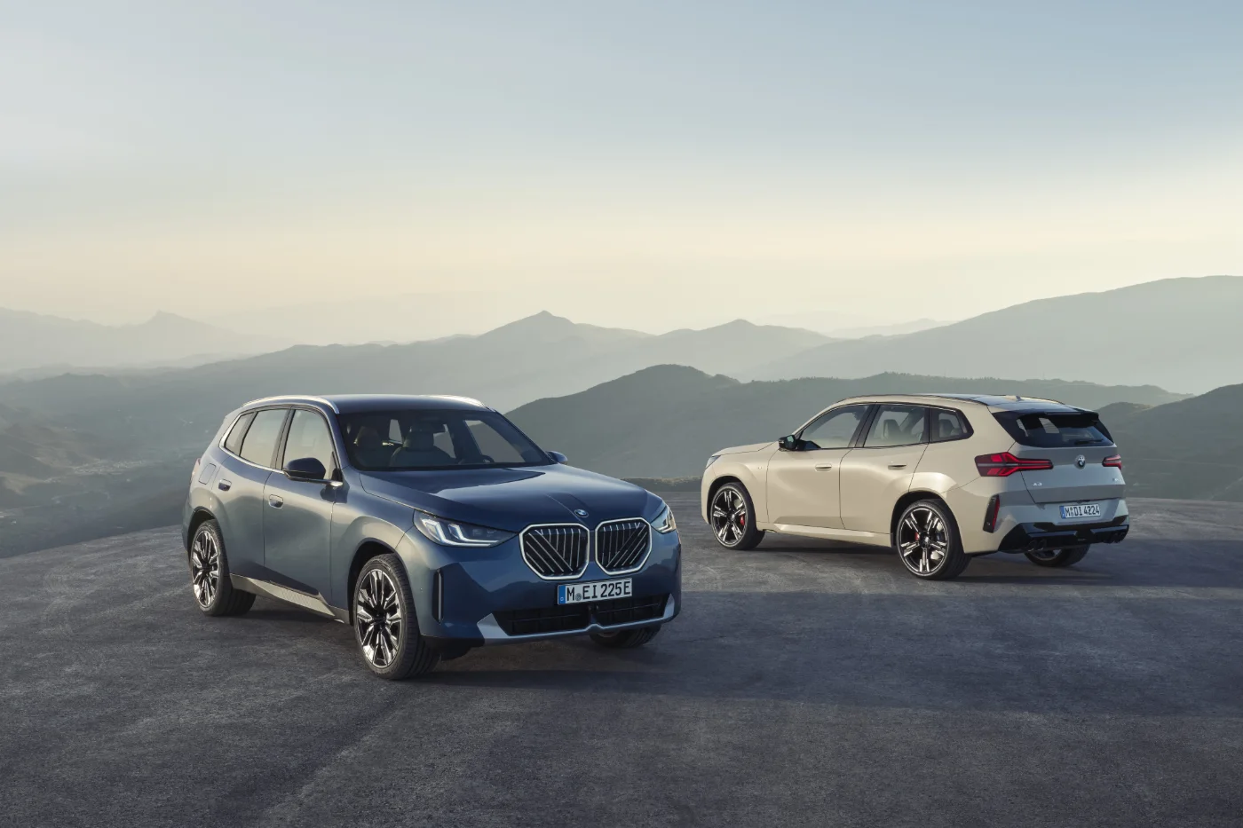 Dwa samochody BMW typu SUV, nowe BMW X3 M50 xDrive, zaparkowane w górskim krajobrazie o świcie. Nowoczesne linie nadwozia i charakterystyczne grille podkreślają sportowy styl i elegancję marki BMW.