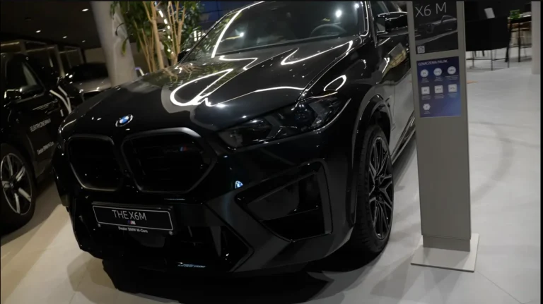Na zdjęciu widzimy nowoczesny, luksusowy samochód BMW X6 M w czarnym, lśniącym lakierze. Auto stoi w eleganckim salonie BMW M-Cars, co potwierdza tabliczka z przodu z napisem THE X6 M – Dealer BMW M-Cars. Reflektory i charakterystyczne, masywne nerki w matowej czerni podkreślają sportowy charakter modelu. Obok pojazdu stoi pionowa tablica informacyjna z oznaczeniami paliw i specyfikacją, a w tle widać inne samochody marki BMW oraz nowoczesne wnętrze salonu – jasną podłogę, rośliny i designerskie oświetlenie odbijające się w karoserii. Całość robi wrażenie mocy, elegancji i prestiżu, a czarny lakier w połączeniu z ciemnymi detalami nadaje samochodowi wyjątkowo agresywny i ekskluzywny wygląd.