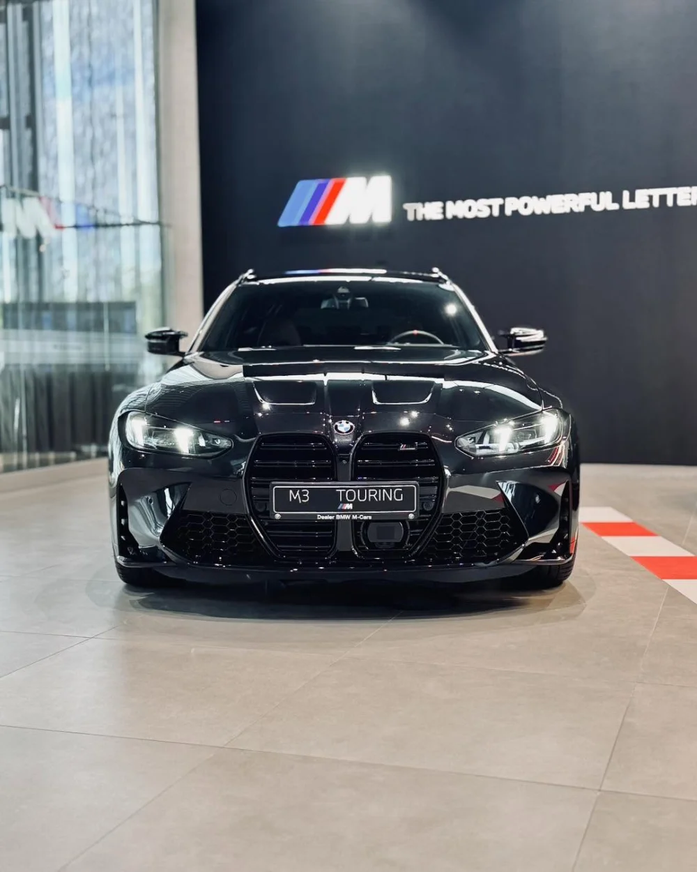 BMW M3 Competition Touring 2026 z przodu w salonie BMW M-Cars, czarny lakier i sportowy pakiet M